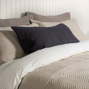 A styled bed with textured beige and taupe bedding, featuring a prominent dark charcoal gray chevron-patterned lumbar pillow in the foreground.